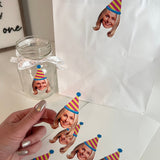 Custom Face Stickers with a womans face cut out wearing a blue, yellow and pink striped party hat and a hand golding a sticker out