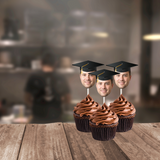 a mans face wearing a graduation cap on lollipop sticks stuck into chocolate cupcakes on a wood table