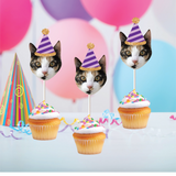 a multicolored cat wearing a purple striped party hat stuck into cupcakes