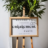 Invitation sign for Sam & Jonah's engagement party on a wooden stand with a plant in the background.