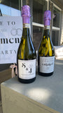 Two bottles of S&J wine with purple corks on a wooden surface, with a blurred background.