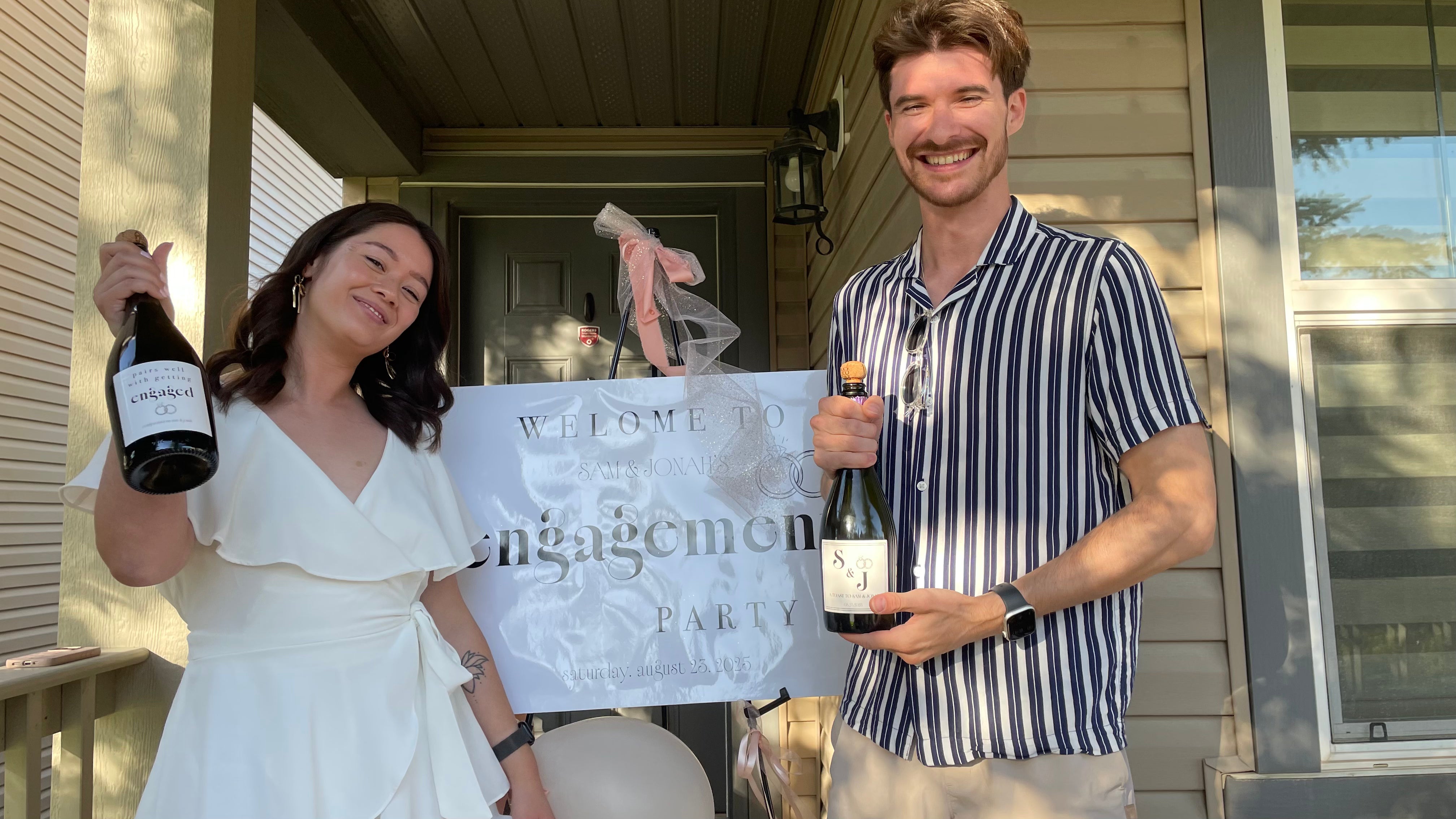 Two people holding bottles of champagne in front of a 'Welcome to our engagement party' sign on a porch.