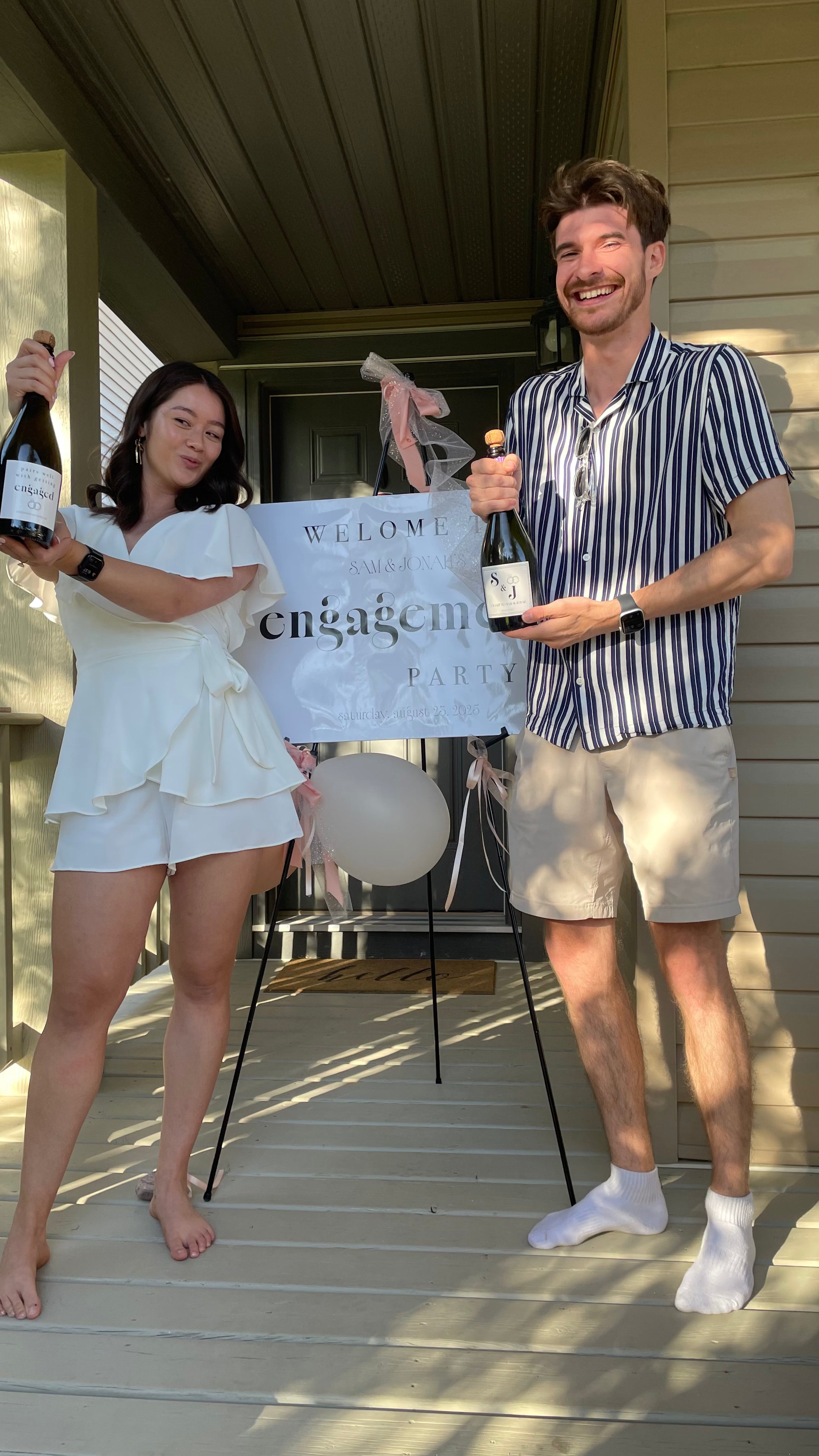 Two people on a porch holding bottles of champagne and an engagement sign.