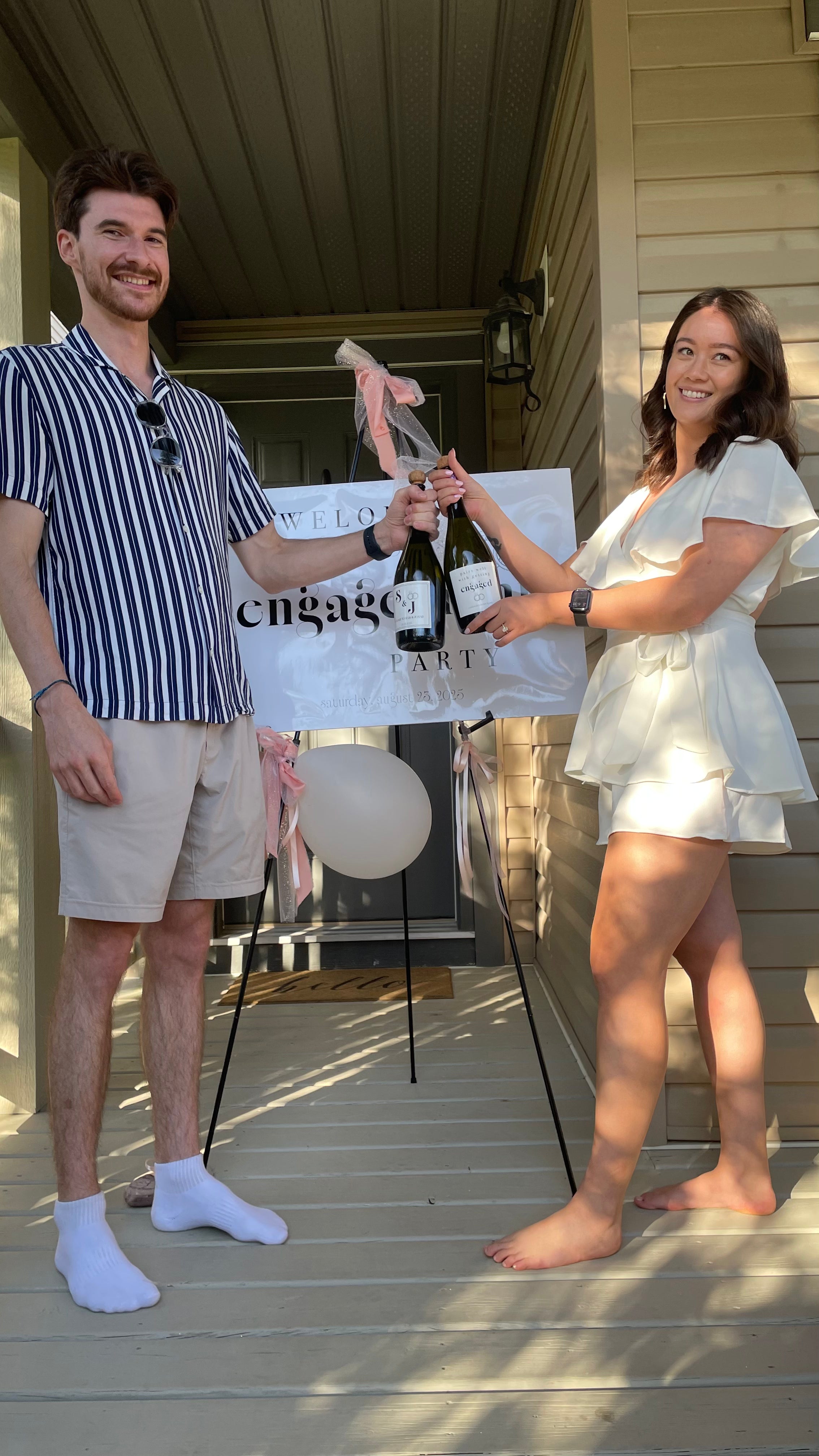 Man and woman holding a bottle of champagne on a porch with 'engage' party sign in the background.