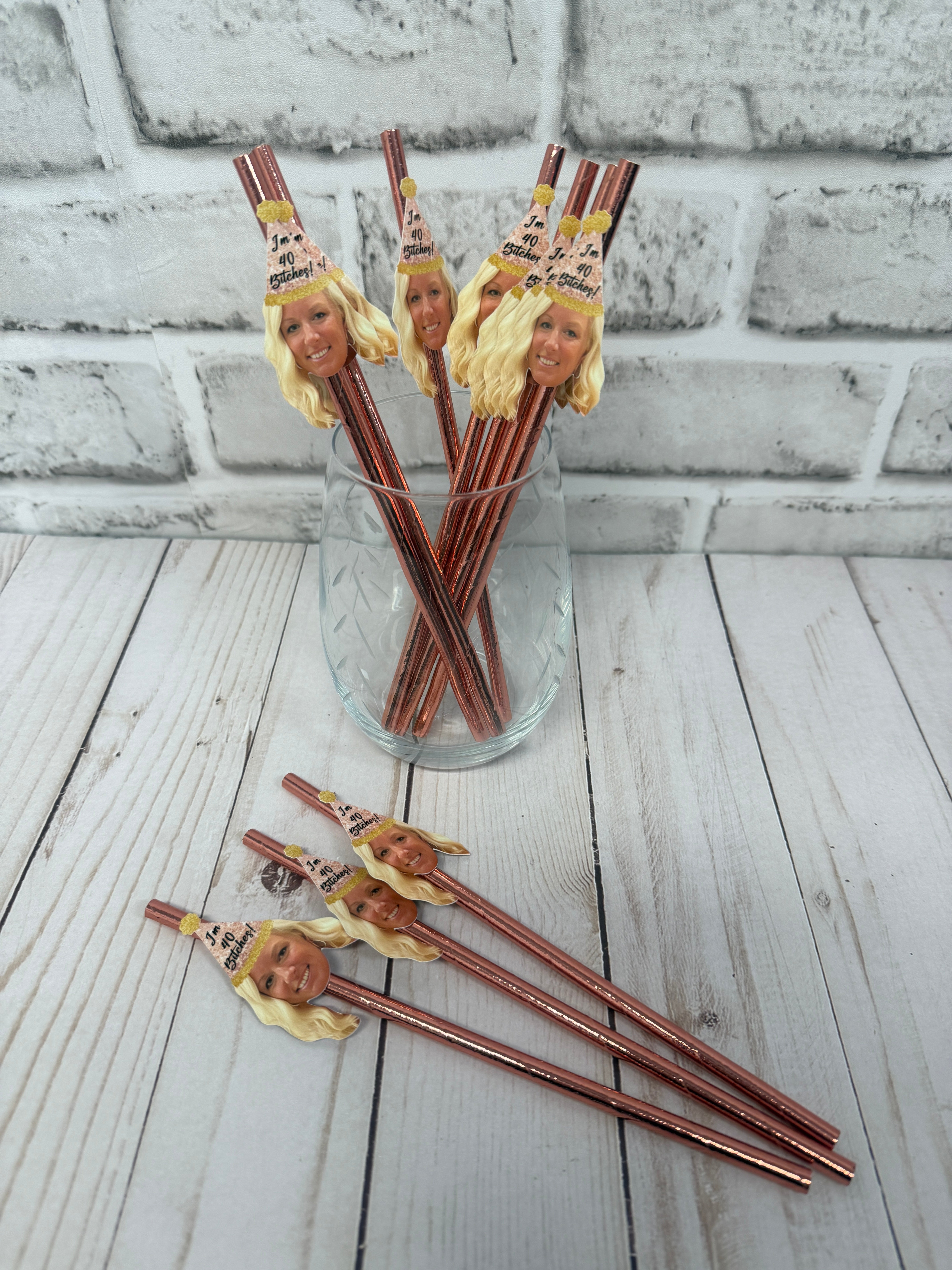 women's face wearing a party hat on rose gold straws in a cup with a light grey brick background