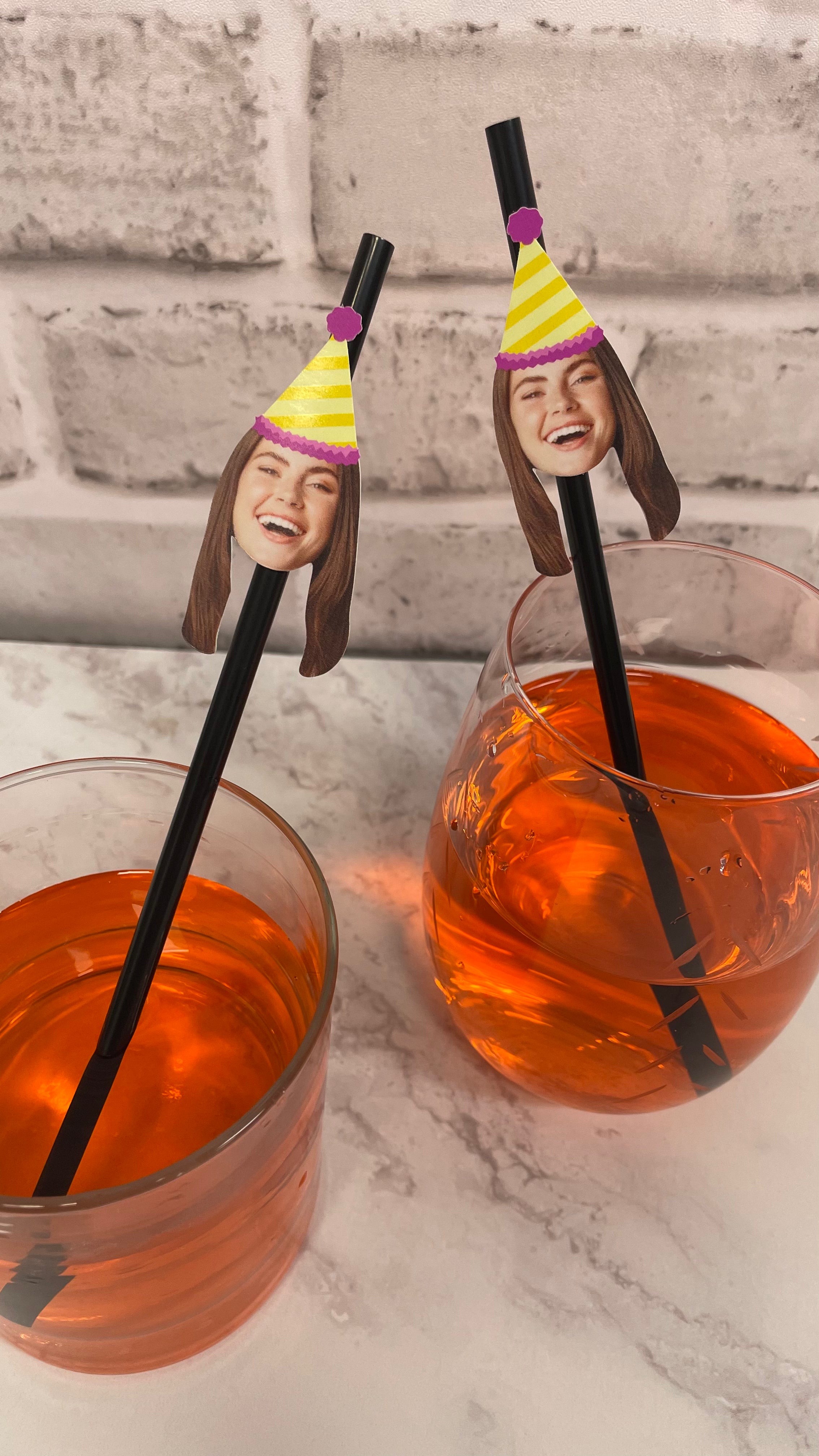 Two glasses of orange liquid with personalized straw toppers on a marble surface.