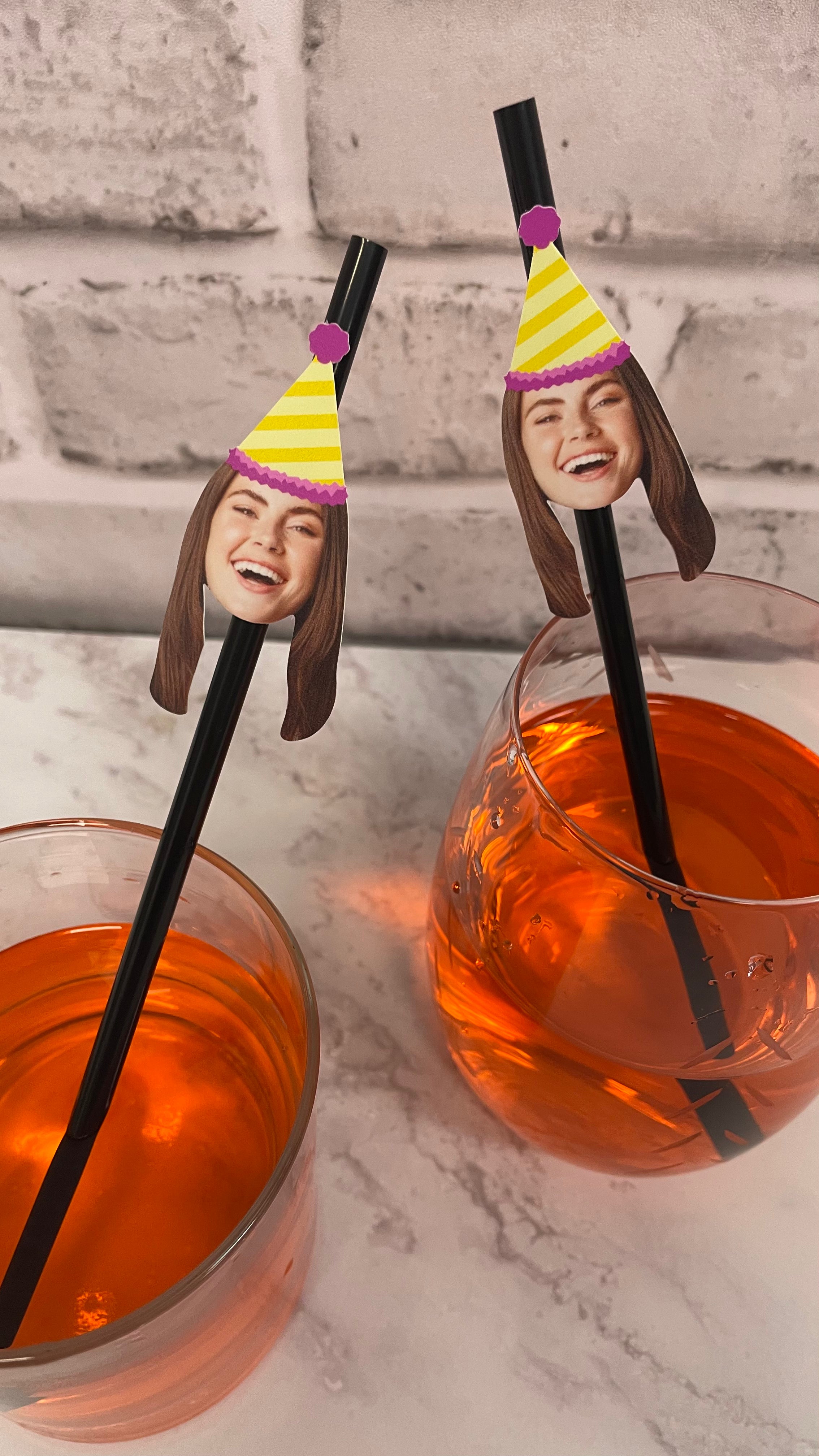 Two cocktail straws with face designs in front of drinks on a marble surface.