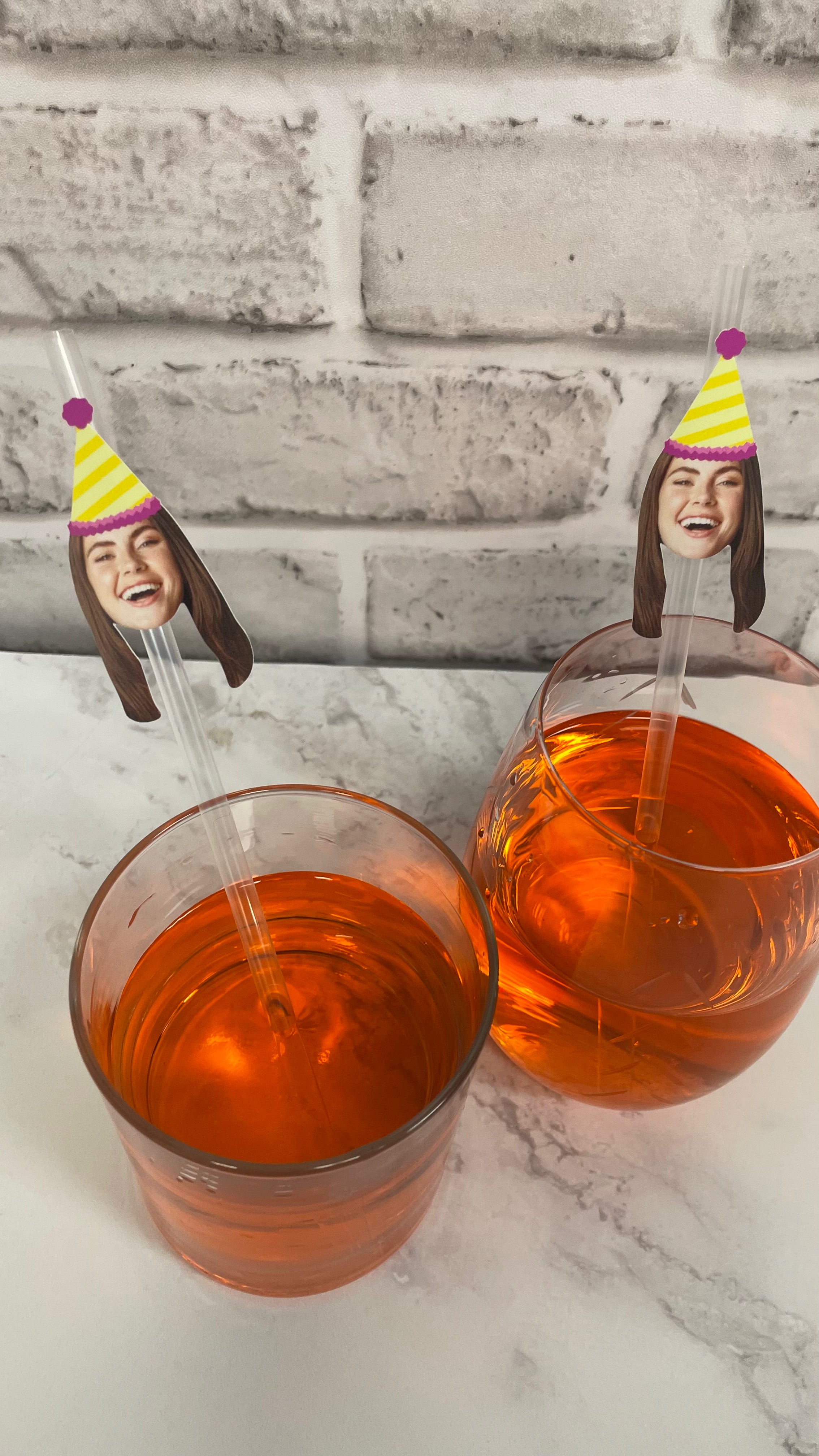 Two glasses of orange liquid with face-shaped straws against a white brick wall background