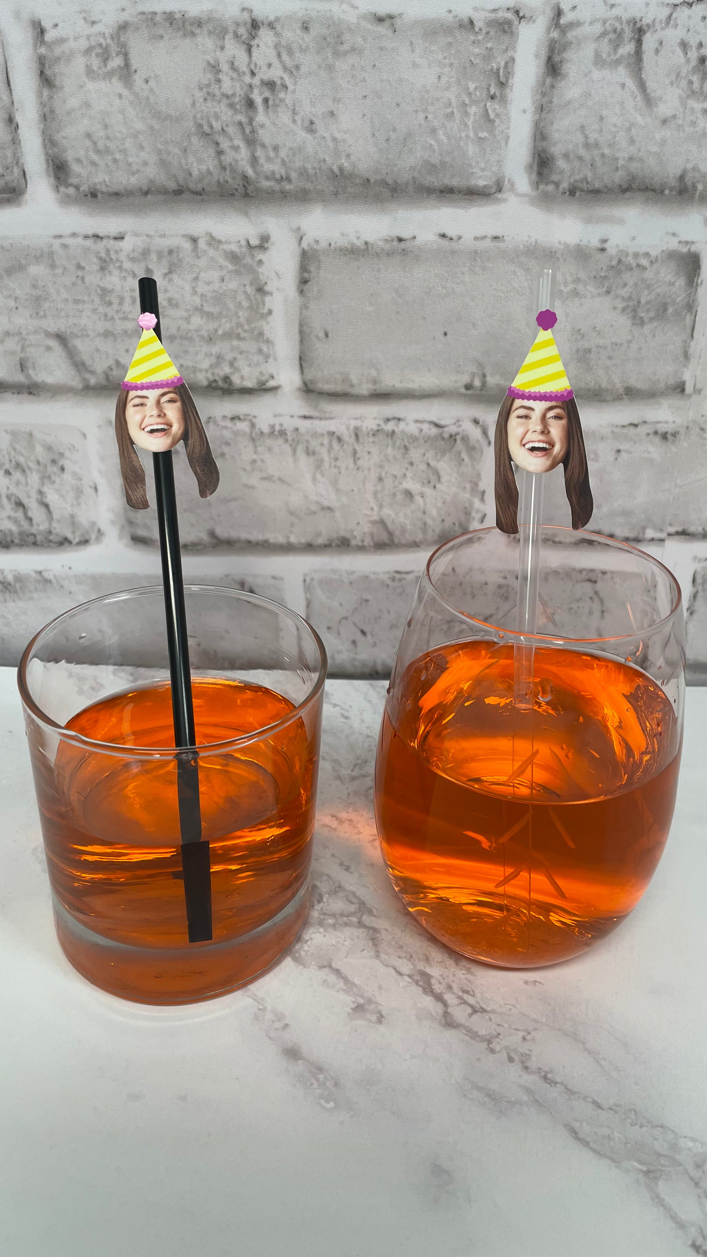 Two glasses of orange liquid with straws featuring faces against a white brick wall.