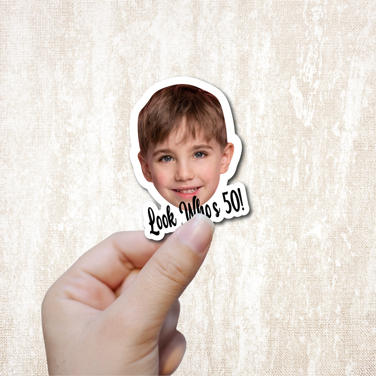 Sticker with a child's face and 'Look Who's 50!' text held by a hand on a textured beige background