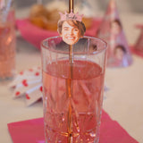Glass with a straw featuring a photo of a smiling person wearing a 'Bride' tiara, on a pink tablecloth.