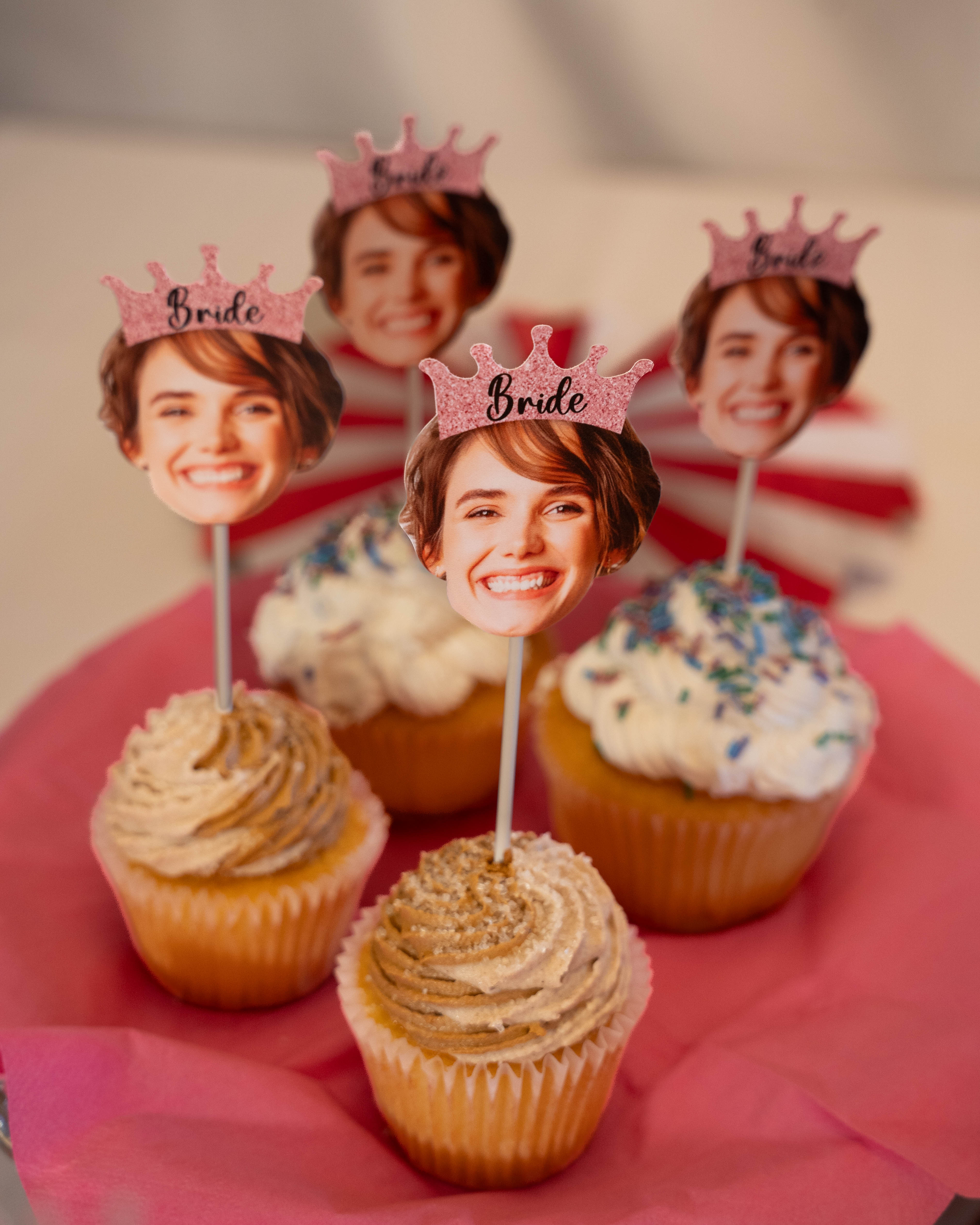 Cupcakes with faces of a bride and groom wearing 'Bride' crowns on a pink surface.