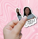 Hand holding a bottle of champagne with a woman in the background on a pink marble patterned background.
