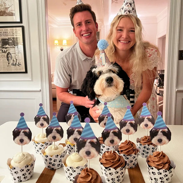 a couple smiling with their dog wearing party hats behind a row of cupcakes featuring cupcake toppers of their dogs face cut out with a blue and purple striped party hat